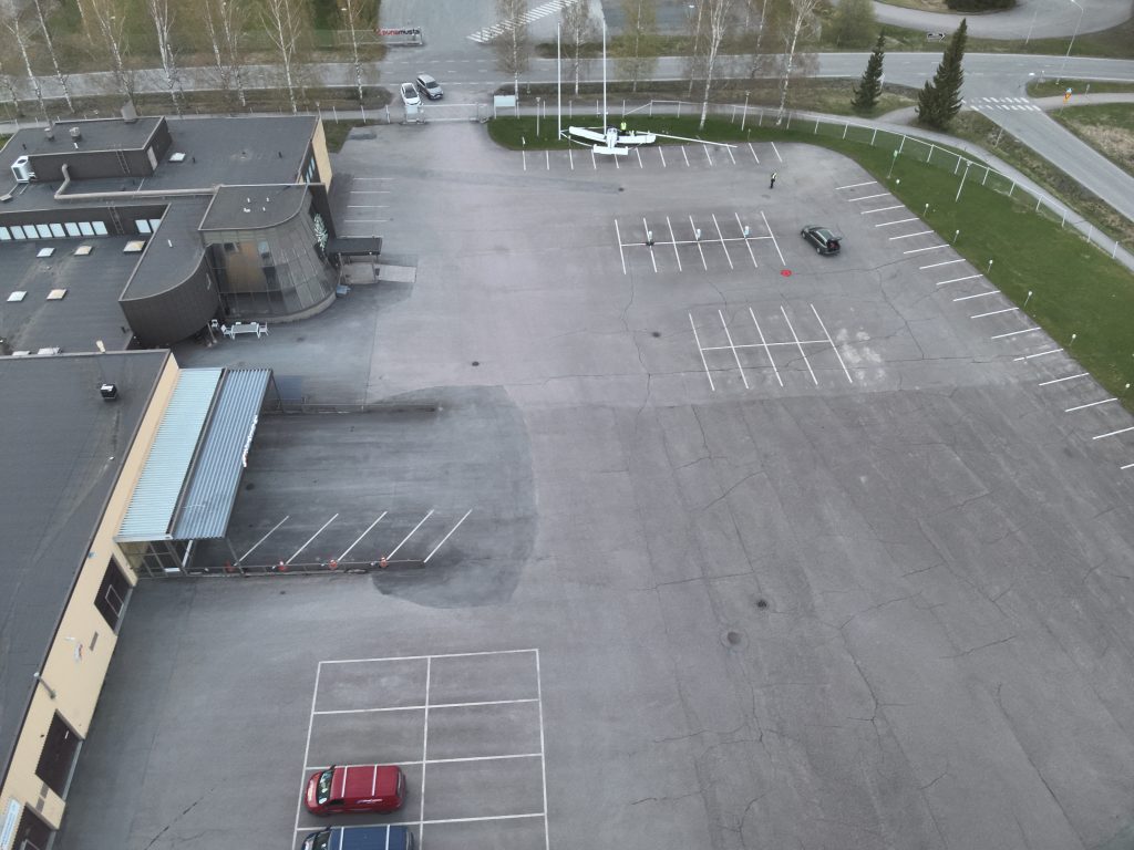 Ilmakuva onnettomuuspaikasta. Etualalla parkkipaikka ja pakkolaskun tehnyt lentokone. Vasemmalla matala teollisuusrakennus ja oikealla autotie.